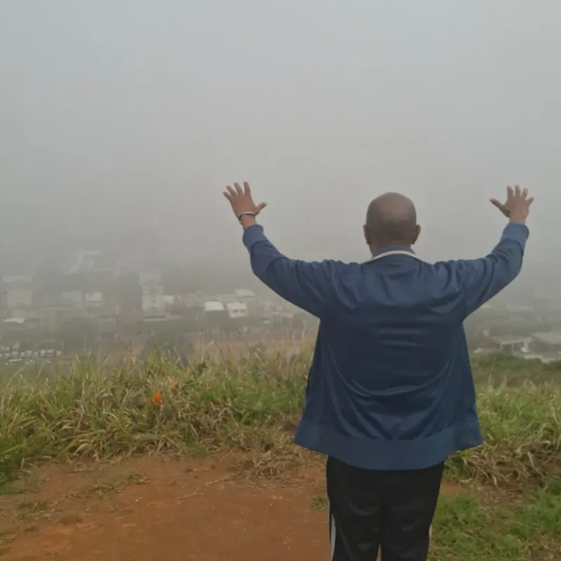 Estevão Maria de costas, com os braços erguidos, no alto do morro coberto de neblina