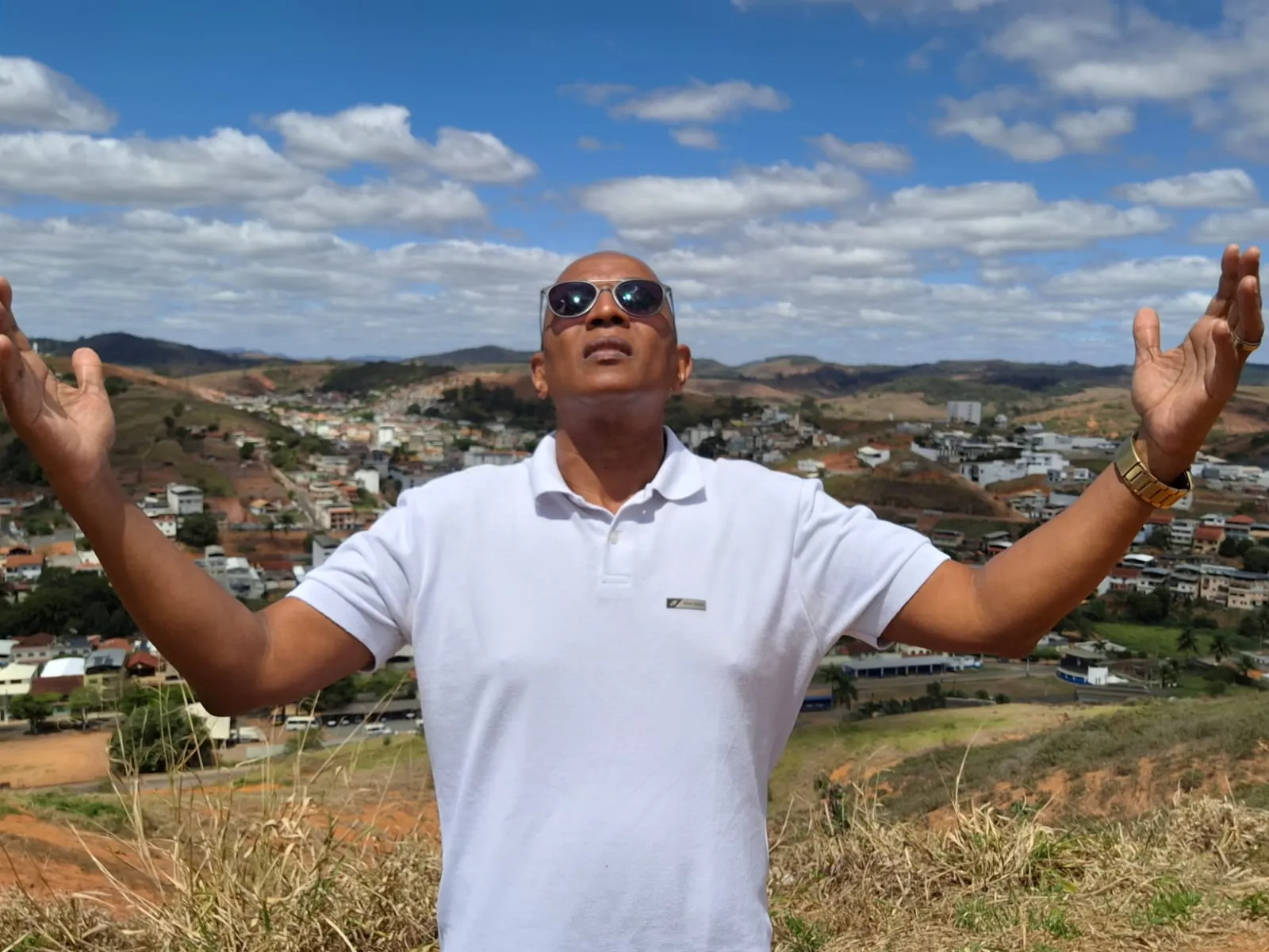 Estevão Maria de braços abertos, olhando para o céu, no alto de um morro com a cidade ao fundo